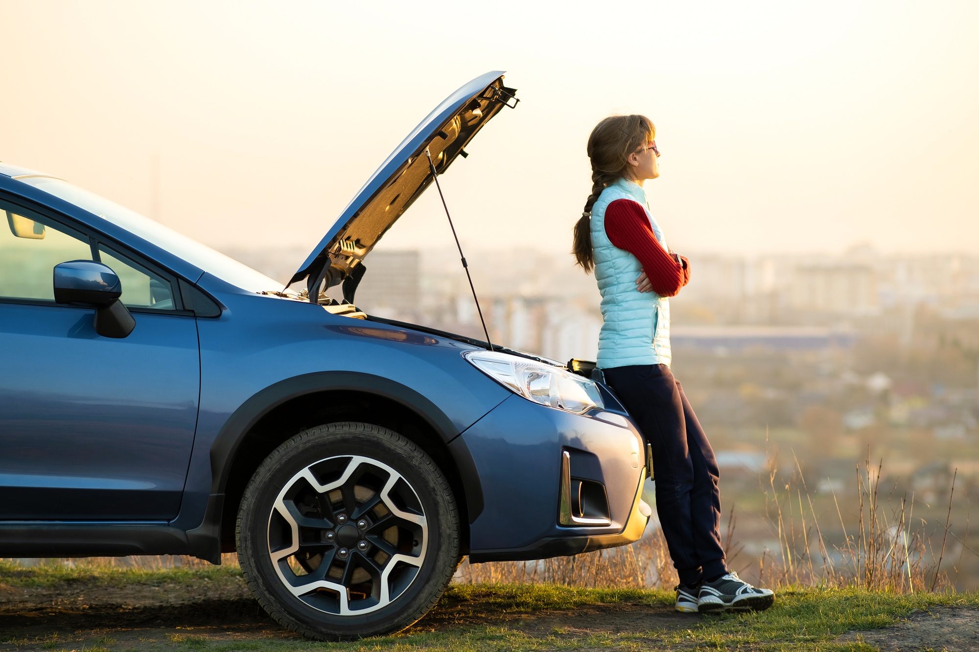 Une jeune femme debout près de la voiture en panne avec un capot sauté ayant des problèmes avec son véhicule. 