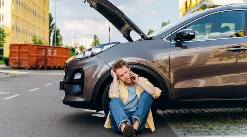 Jeune homme utilisant un téléphone portable près d'une voiture cassée dans la ville.